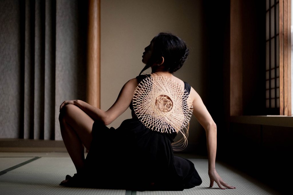 A dark room with a light shining down on to a sitting figure, back to the camera dressed in black with a light intricate bamboo dressing on the back.