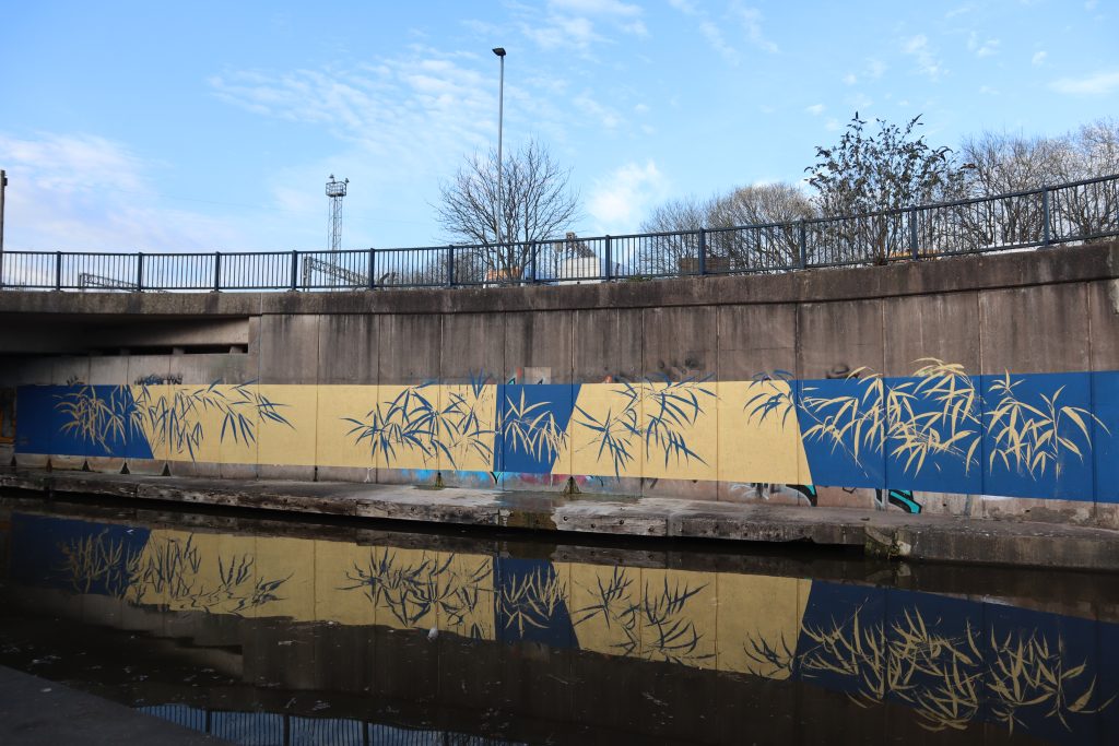 A gold and blue long painted mural on a concrete structure alongside the canal. With gold and blue bamboo painted over the opposite colour for contrast.