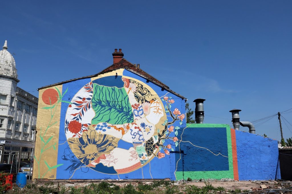 A large gable end with gold blue, terracotta green colours to make a circle motif. A bamboo is growing over a gold panel on the left and the other botanical patterns make up a kintsugi style plate of Spode patterns.